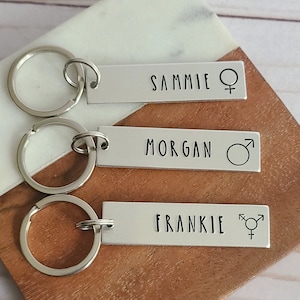 May include: Three silver keychains with personalized name tags. The top tag reads "SAMMIE" with a female symbol. The middle tag reads "MORGAN" with a male symbol. The bottom tag reads "FRANKIE" with a transgender symbol.