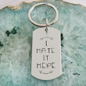 May include: Silver keychain with a rectangular tag that says "I hate it here" with arrows on either side. The keychain is on a green and gold agate coaster.