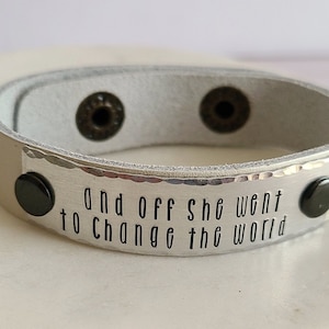 May include: A white leather bracelet with silver accents and black snaps. The bracelet has the text "And off she went to change the world" engraved on it.
