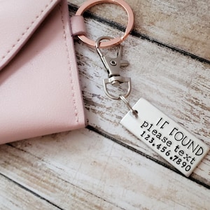 May include: A blush pink wallet with a rose gold snap closure and a matching key ring. Attached is a small metal tag with the text "IF FOUND please text 123.456.7890". The wallet and key ring are on a wooden surface.