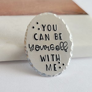 May include: Oval-shaped, hammered metal pendant with the phrase "YOU CAN BE yourself WITH ME" in black lettering. The pendant has a textured surface and is set against a white and brown background.