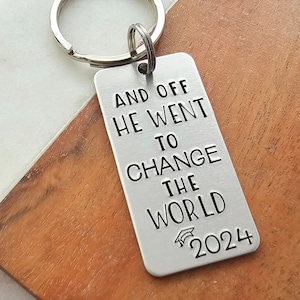 May include: Silver keychain with the text "And off he went to change the world 2024" and a graduation cap icon.