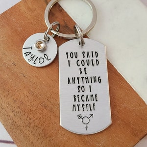 May include: Silver keychain with a tag that reads "You said I could be anything so I became myself" with a transgender symbol. The keychain also has a smaller tag with the name "Taylor" and a small clear rhinestone.