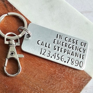 May include: A silver metal keychain with a rectangular tag. The tag has the text "IN CASE OF EMERGENCY CALL STEPHANIE 123.456.7890". The keychain is on a wooden and marble surface.