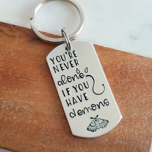May include: Silver keychain with a silver tag that reads "You're never alone if you have demons" with a moth design.