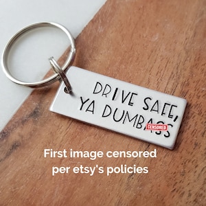 May include: A silver keychain with a rectangular tag that reads "Drive Safe, Ya Dumb[CENSORED]" in black lettering. The keychain is attached to a silver key ring.