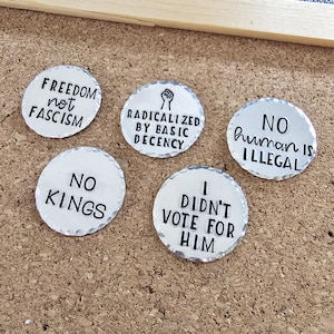 May include: Five small, round, silver-colored metal pins with black text. The pins have various phrases: "FREEDOM not FASCISM", "RADICALIZED BY BASIC DECENCY", "NO human is ILLEGAL", "NO KINGS", and "I DIDN'T VOTE FOR HIM".