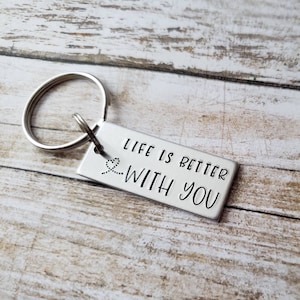 May include: A silver rectangular keychain with the phrase "LIFE IS BETTER WITH YOU" stamped in black. The keychain has a silver ring for attaching keys. The background is a light wood surface.