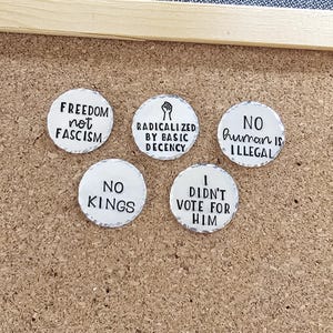 Pode incluir: Cinco bot&otilde;es redondos brancos com texto preto numa placa de corti&ccedil;a. Os bot&otilde;es apresentam v&aacute;rias frases: "FREEDOM not FASCISM", "RADICALIZED BY BASIC DECENCY", "NO human is ILLEGAL", "NO KINGS" e "I DIDN'T VOTE FOR HIM".