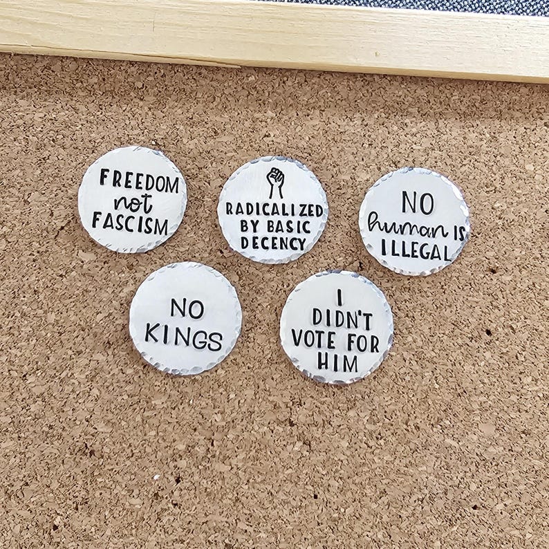 Pode incluir: Cinco bot&otilde;es redondos brancos com texto preto numa placa de corti&ccedil;a. Os bot&otilde;es apresentam v&aacute;rias frases: "FREEDOM not FASCISM", "RADICALIZED BY BASIC DECENCY", "NO human is ILLEGAL", "NO KINGS" e "I DIDN'T VOTE FOR HIM".