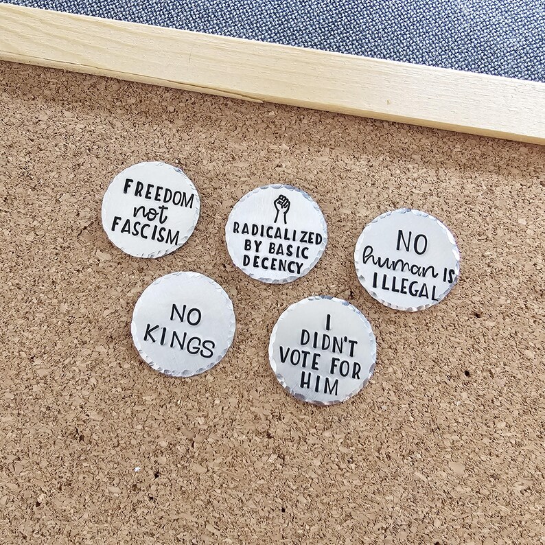 Pode incluir: Cinco bot&otilde;es redondos prateados com texto preto. Os bot&otilde;es apresentam frases como "Freedom not Fascism", "Radicalized by Basic Decency" e "No Human is Illegal". Est&atilde;o dispostos numa placa de corti&ccedil;a.