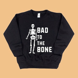 Pu&ograve; includere: Felpa nera con un grafico di scheletro bianco e il testo "Bad to the Bone".