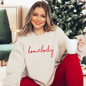 Puede incluir: Una sudadera de color crema con la palabra "homebody" escrita en cursiva roja.