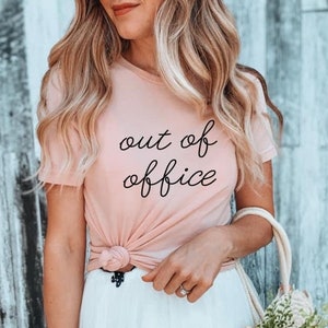 Pode incluir: Uma mulher vestindo uma camiseta rosa com o texto "out of office" em escrita preta. Ela est&aacute; usando uma saia branca e &oacute;culos de sol. Ela est&aacute; segurando uma bolsa de palha com flores.