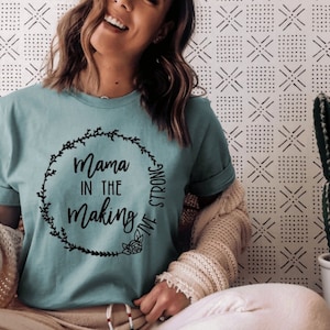 May include: A teal blue t-shirt with a black graphic that says "Mama in the Making" inside a wreath. The wreath has a black ribbon that says "Five Strong" on it.