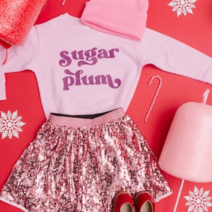 Pu&ograve; includere: Gonna di paillettes rosa con cintura nera e una felpa rosa con la scritta "sugar plum" in lettere viola.