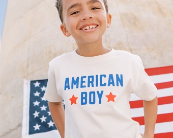Può includere: Un bambino piccolo con una maglietta bianca con il testo "AMERICAN BOY" in lettere blu e due stelle rosse. È in piedi davanti a una bandiera americana.