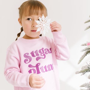 Pu&ograve; includere: Una bambina indossa una felpa rosa con la scritta "Sugar Plum" in lettere viola. Sta tenendo un ornamento a forma di fiocco di neve bianco davanti al viso.