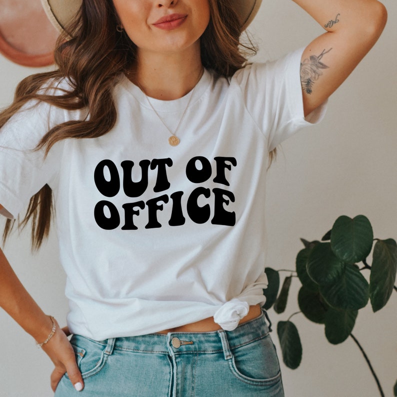 Op de afbeelding: Wit T-shirt met zwarte tekst die "OUT OF OFFICE" luidt in een retro lettertype.