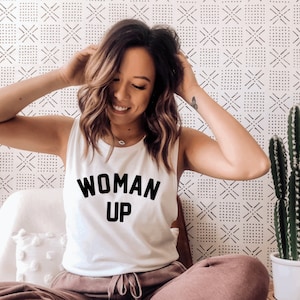 May include: A white tank top with the text "WOMAN UP" printed in black. The model is wearing the tank top and smiling.