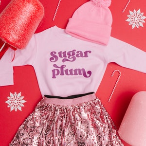 Pu&ograve; includere: Gonna di paillettes rosa con cintura nera, felpa rosa con il testo "sugar plum" in viola, berretto rosa con pon pon bianco e sfondo rosso con fiocchi di neve bianchi e bastoncini di zucchero.