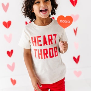 May include: A child wearing a white t-shirt with the words "HEART THROB" printed in red. The child is holding a red heart-shaped sign that says "XOXO".