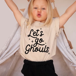 May include: A child wearing a beige t-shirt with the text "Let's go Ghouls" in black. The child is wearing black pants and has blonde hair.