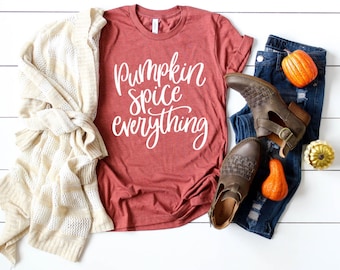 May include: A rust-colored t-shirt with the text "Pumpkin Spice Everything" in white lettering. The shirt is laid out on a white wooden surface with a white cardigan, blue jeans, and small orange pumpkins.