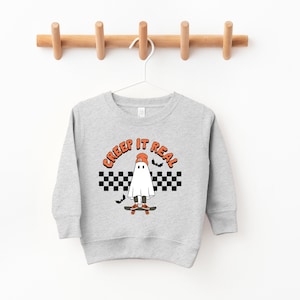 May include: A gray sweatshirt with a ghost wearing a hat and skateboarding. The text "CREEP IT REAL" is above the ghost. The ghost is on a checkered background with bats.