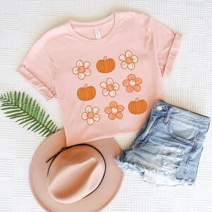 May include: A pink t-shirt with a graphic design of orange pumpkins and orange flowers. The shirt is laid flat on a white surface with a brown hat and blue denim shorts.