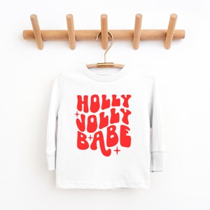 May include: White long-sleeve shirt with a red graphic that says "HOLLY JOLLY BABE" in a retro font.