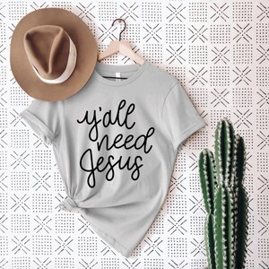 May include: A gray t-shirt with the text "y'all need Jesus" written in black cursive lettering.
