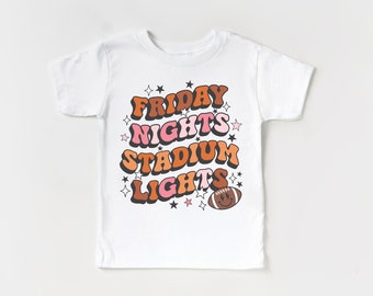 May include: White t-shirt with a retro-style graphic design that says "FRIDAY NIGHTS STADIUM LIGHTS" with a football graphic. The text is in orange, pink, and brown.