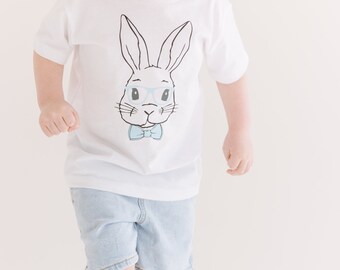 Può includere: Una maglietta bianca con un'illustrazione in bianco e nero di un coniglio con occhiali blu e un papillon.