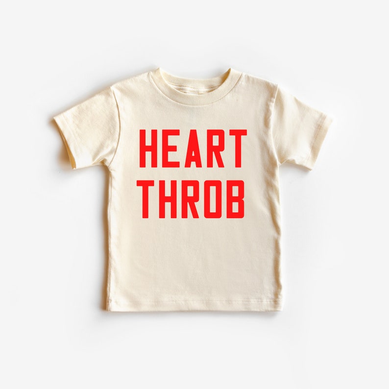 May include: A cream colored t-shirt with short sleeves and the words "HEART THROB" printed in red.