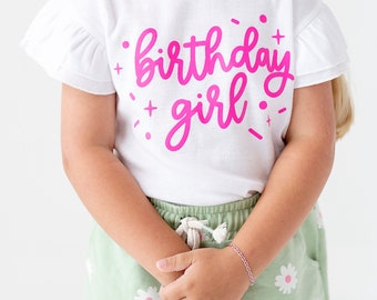 May include: A white t-shirt with pink lettering that says "birthday girl" with pink confetti around the text. The shirt has ruffled sleeves and is being worn by a child.