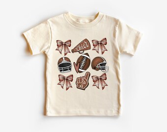 May include: A cream colored t-shirt with brown football themed graphics. The graphics include a football, a helmet, a cheerleader's megaphone, a heart, a number one hand gesture, and bows.
