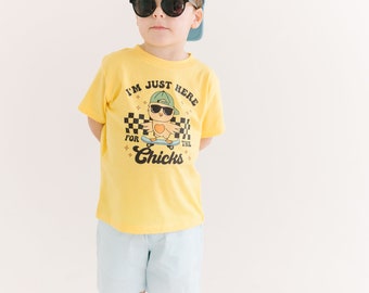 Può includere: Una maglietta gialla con un pulcino dei cartoni animati che indossa occhiali da sole e un cappello, mentre fa skateboard. Il testo sulla maglietta recita "I'm just here for the chicks".