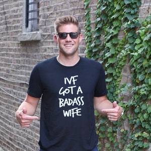 May include: A man wearing sunglasses is standing in front of a brick wall with ivy growing on it. He is wearing a black t-shirt with the text "IVF GOT A BADASS WIFE" printed in white.