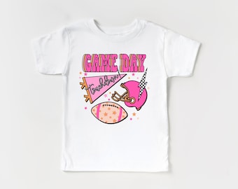 May include: White t-shirt with a pink "GAME DAY" graphic. The graphic includes a pink football helmet, a pink pennant with the word "Touchdown!" and a football with orange and white stars.