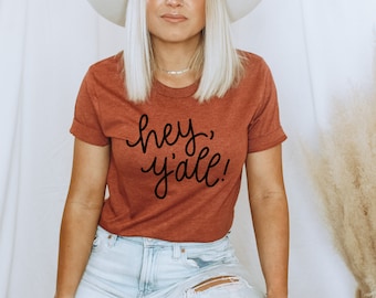 May include: Rust-colored t-shirt with rolled sleeves and the phrase "hey, y'all!" in black script. The shirt is paired with light-wash jeans. A white hat and silver necklace complete the look. The overall style is casual and relaxed.