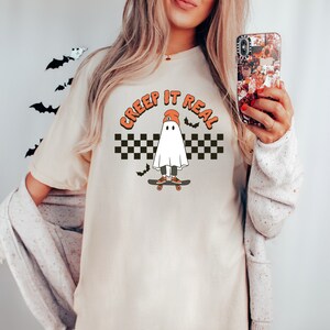 May include: A white t-shirt with a graphic of a ghost wearing a beanie and skateboarding. The text "CREEP IT REAL" is written in a circle around the ghost.