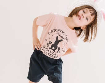 Può includere: Una giovane persona indossa una maglietta rosa con un coniglio nero e il testo "Cottontail Candy Co. Premium 1923 St. Chocola & Jellybeans" stampato sul davanti. Indossa pantaloncini neri e un cappello bianco con una fascia per capelli con orecchie di coniglio rosa.