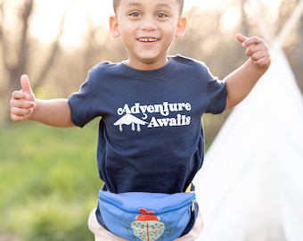 May include: A young child wearing a navy blue t-shirt with the text "Adventure Awaits" and a white mountain graphic. The child is also wearing a blue fanny pack with a ladybug design.