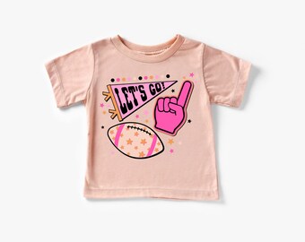 May include: A pink t-shirt with a graphic design featuring a pink pennant with the text "LET'S GO!" and a pink hand with a raised index finger. The graphic also includes a football with orange and white stars.