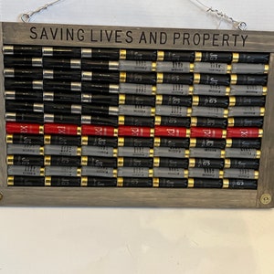May include: A wooden framed American flag made of shotgun shells. The flag is displayed on a white wall. The frame is gray and has the words "SAVING LIVES AND PROPERTY" written on it.