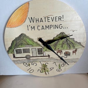 May include: Round wooden clock with a camping theme. The clock face features a hand-drawn illustration of a camper, mountains, and the text "WHATEVER! I'M CAMPING...". The clock hands are black.