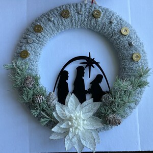 May include: A gray knitted wreath decorated with gold bullet casings and a white poinsettia. A black silhouette of the nativity scene is centered on the wreath.