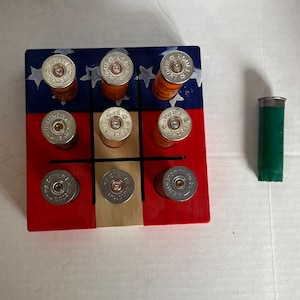 May include: A wooden game board with a red, white, and blue American flag design. The board has nine spaces, each containing a shotgun shell. The shells are arranged in a tic-tac-toe pattern. A green shotgun shell is also visible in the image.