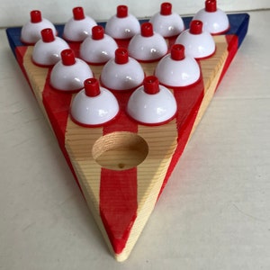 May include: A wooden triangle game board with red and white stripes painted on it. There are 18 white plastic cups with red tops, arranged in a triangular pattern on the board.